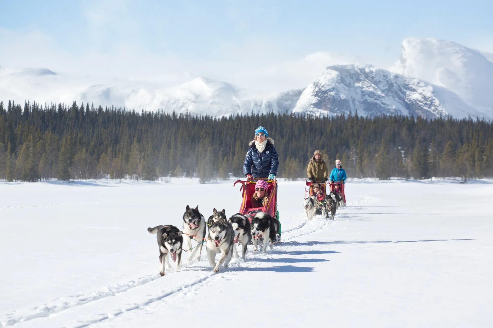 A husky team pulls a sled with two people across a snowfield, more sleds behind and snowy mountains in the distance.