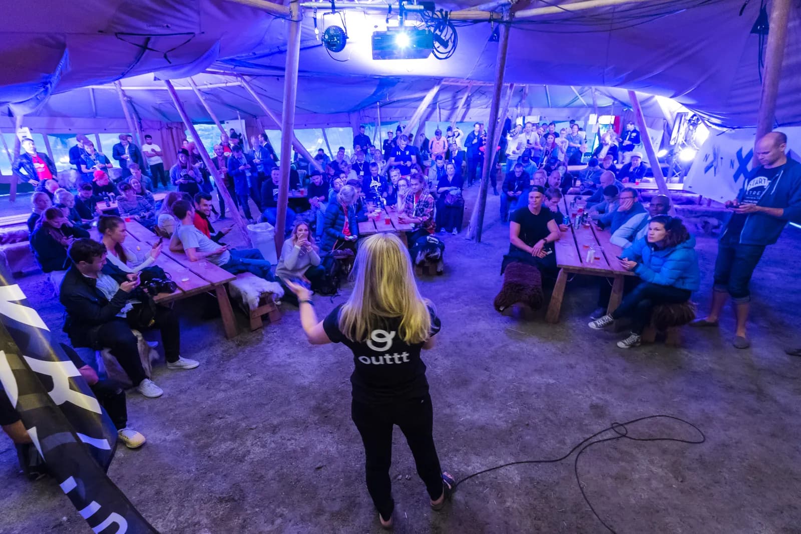 A woman with a microphone talks to a full, purple-lit tipi tent with long wooden tables and sheepskins.