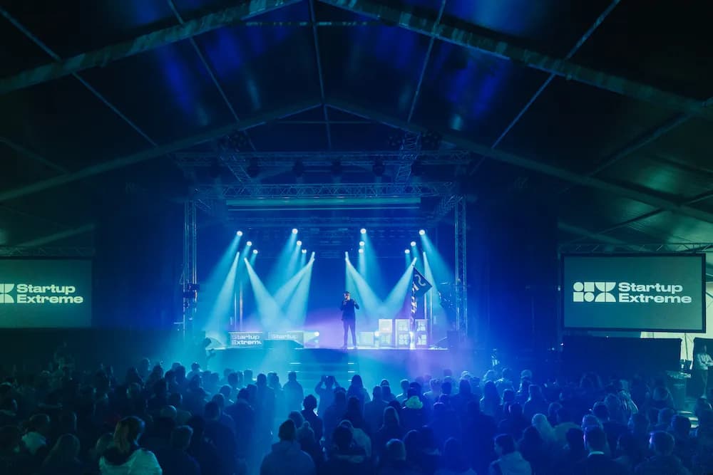 A packed tent in blue stage light, a speaker between two Startup Extreme screens, seen from the back of the crowd in Voss.