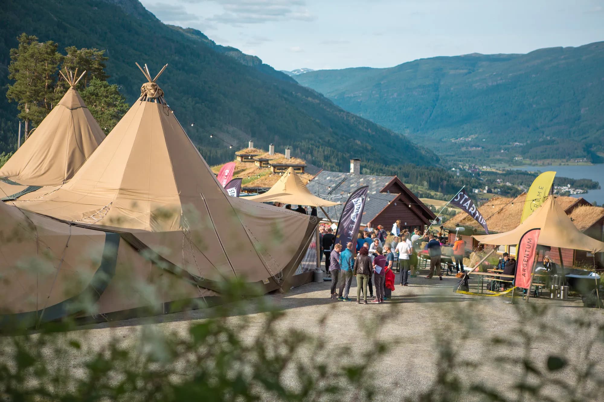 The tipi camp in a green valley at Voss, with flags and people mingling and mountains behind.