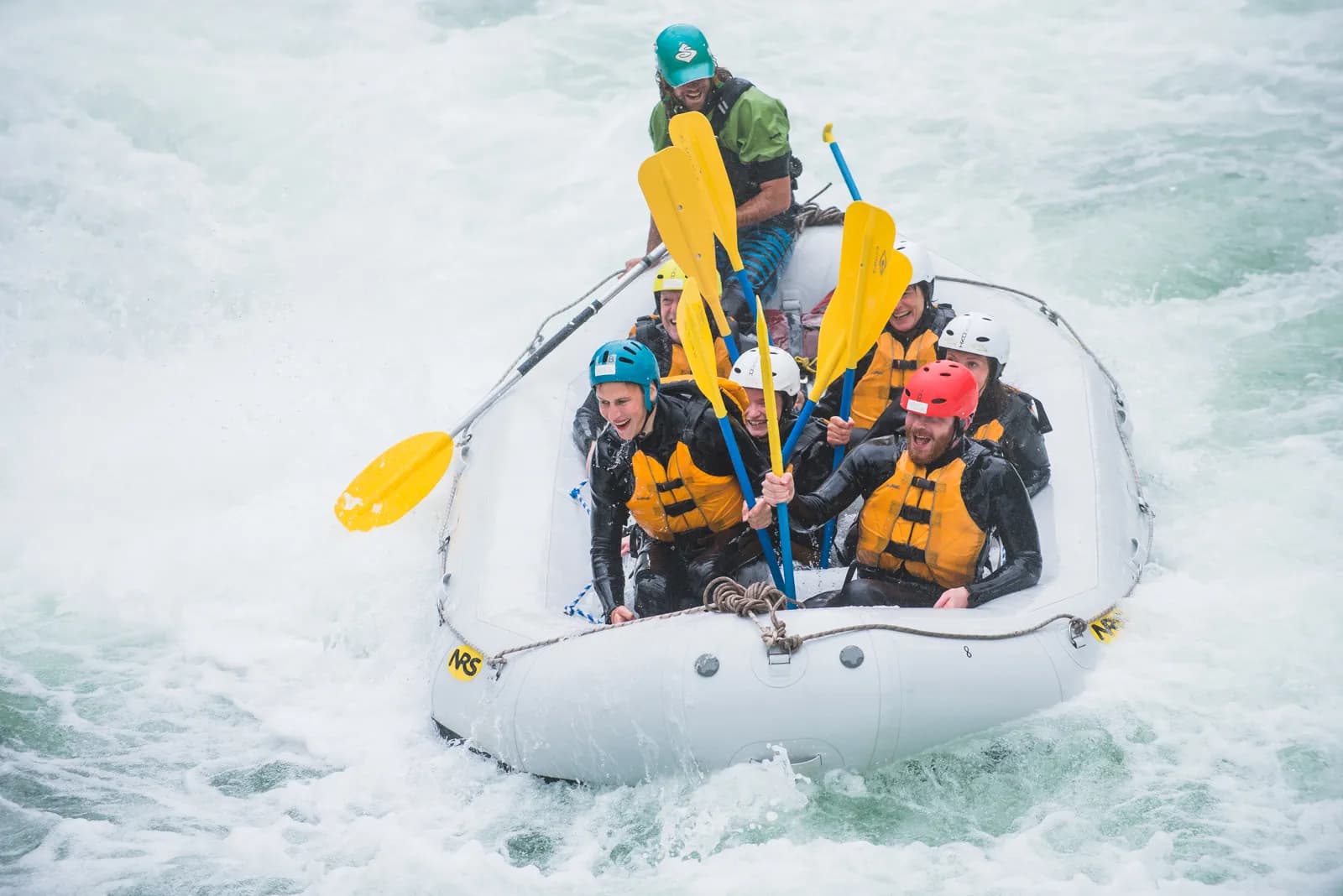 A raft crew in helmets and life jackets paddles through heavy white water.
