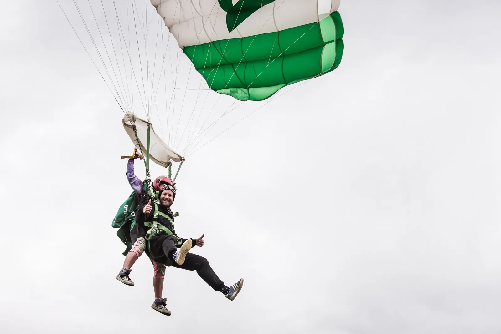 A tandem skydiver under a green and white canopy, the passenger smiling with a thumbs up.