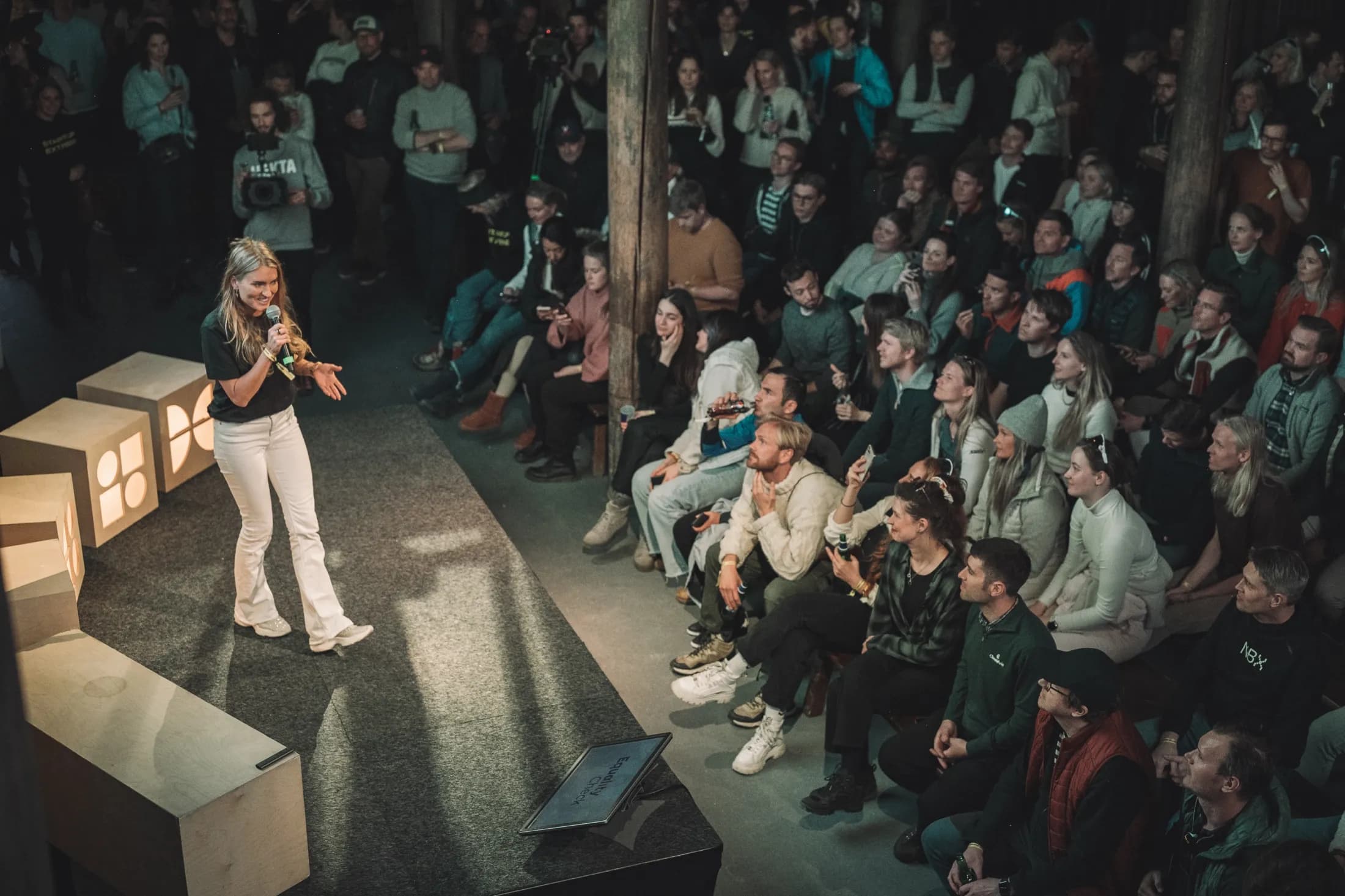 A speaker on a low stage in front of a tightly packed crowd in the dark timber hall at Stavkroa.
