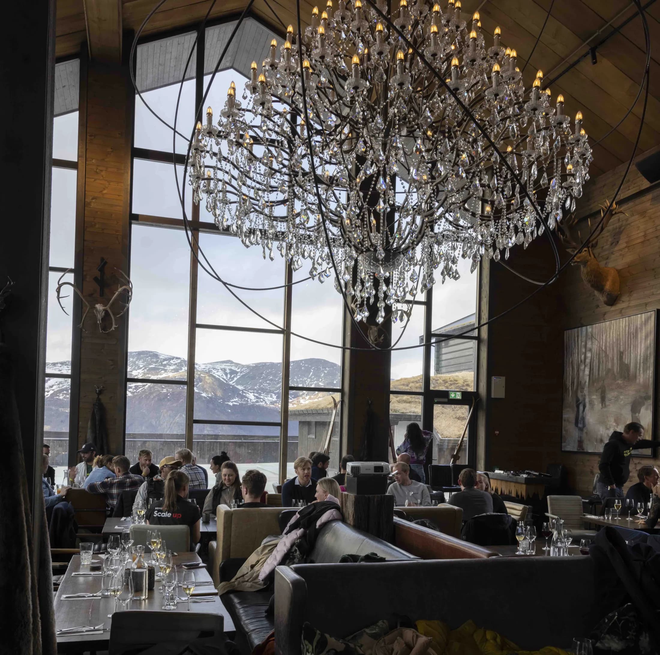 Guests at dinner tables under a large crystal chandelier, panorama windows facing the mountain.