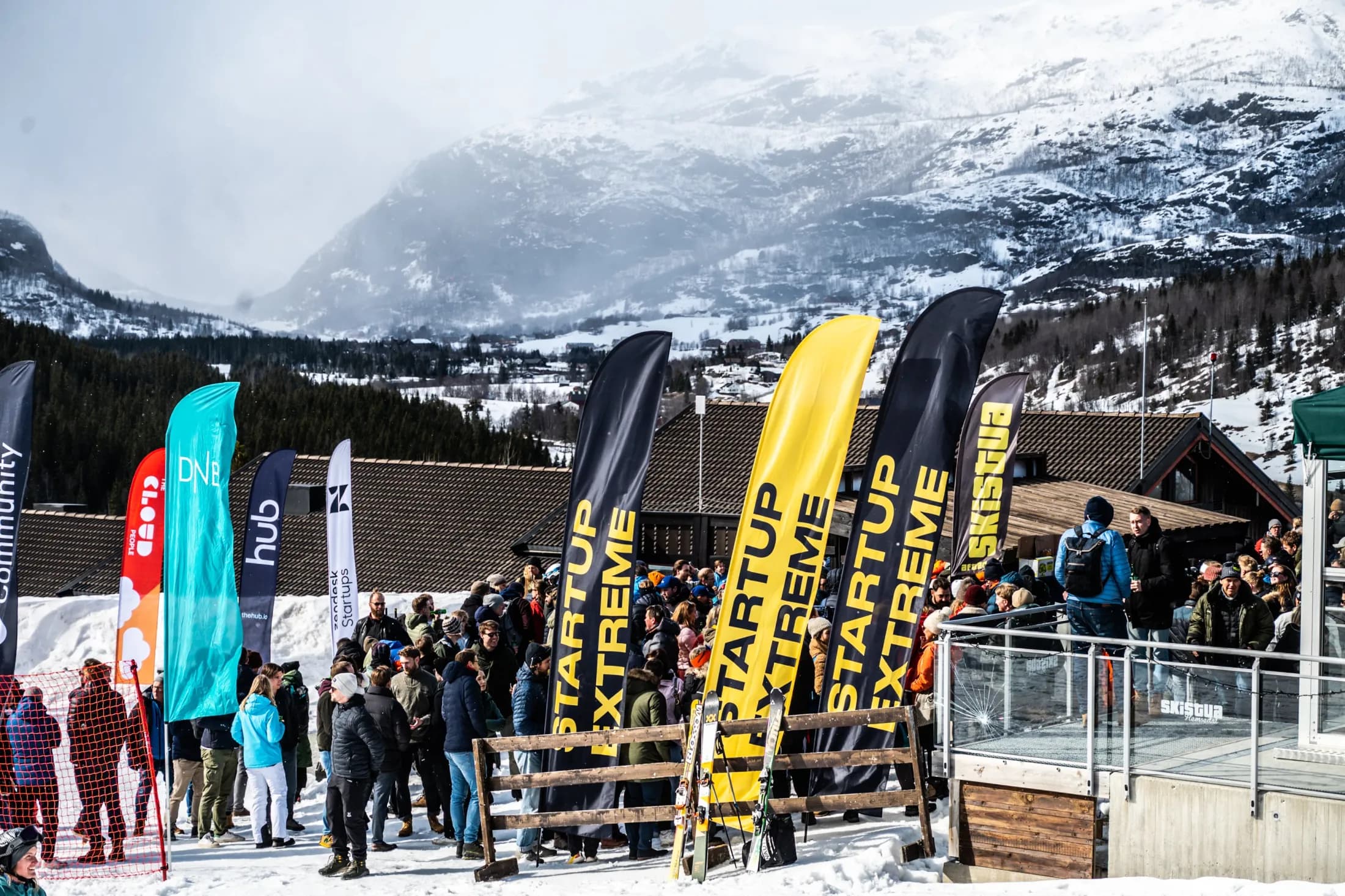 A crowd outside Stavkroa among yellow Startup Extreme beach flags, snow-covered mountains behind.