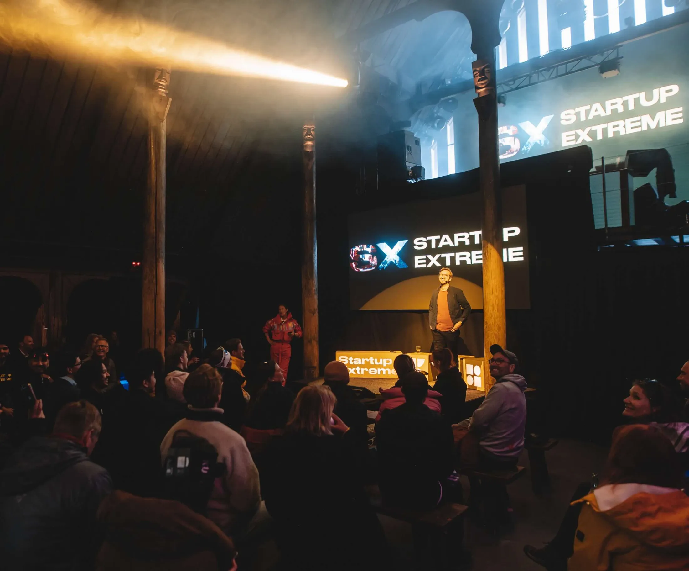 A speaker on the main stage in a dark timber hall, a beam of light above and the Startup Extreme screen behind.