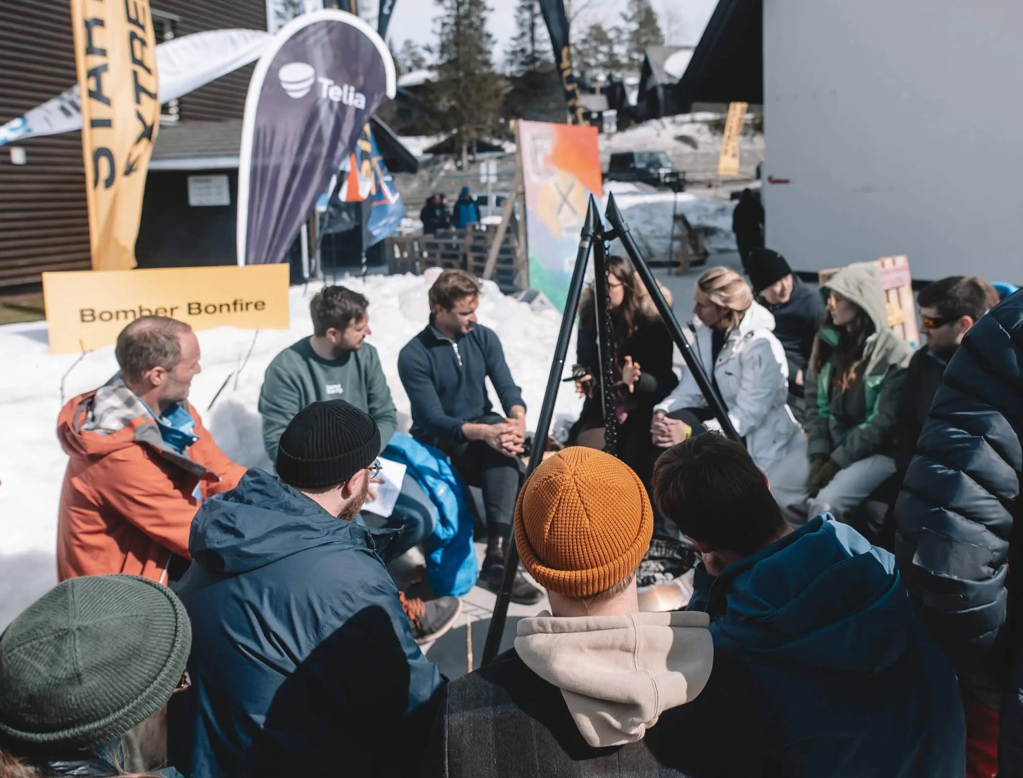 Around twenty people in winter jackets and beanies sit in a tight circle around a fire pan in the snow, next to a sign reading Bomber Bonfire.