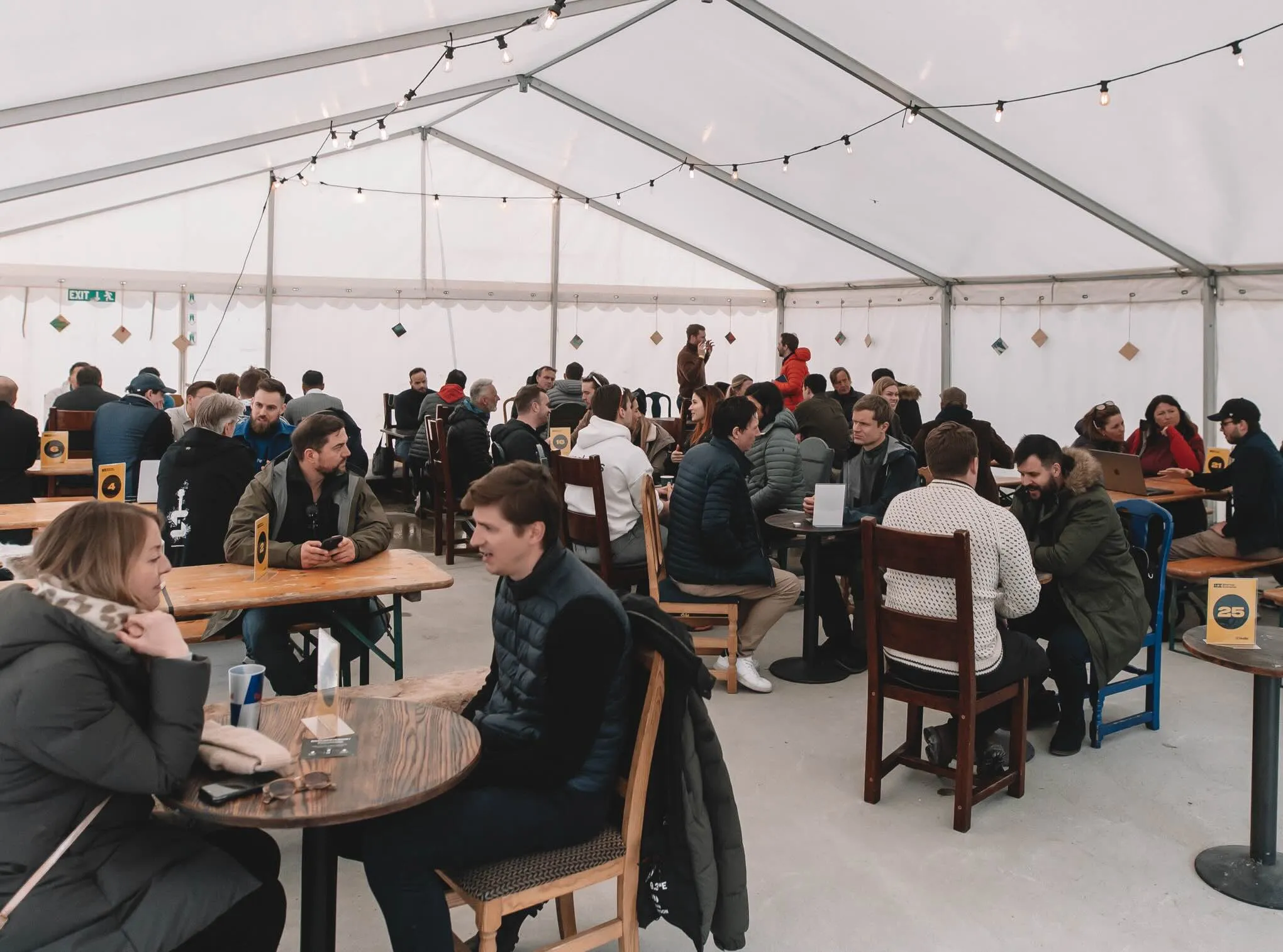 A white tent with string lights full of small tables, people in jackets in one-to-one conversations, yellow table numbers on each table.