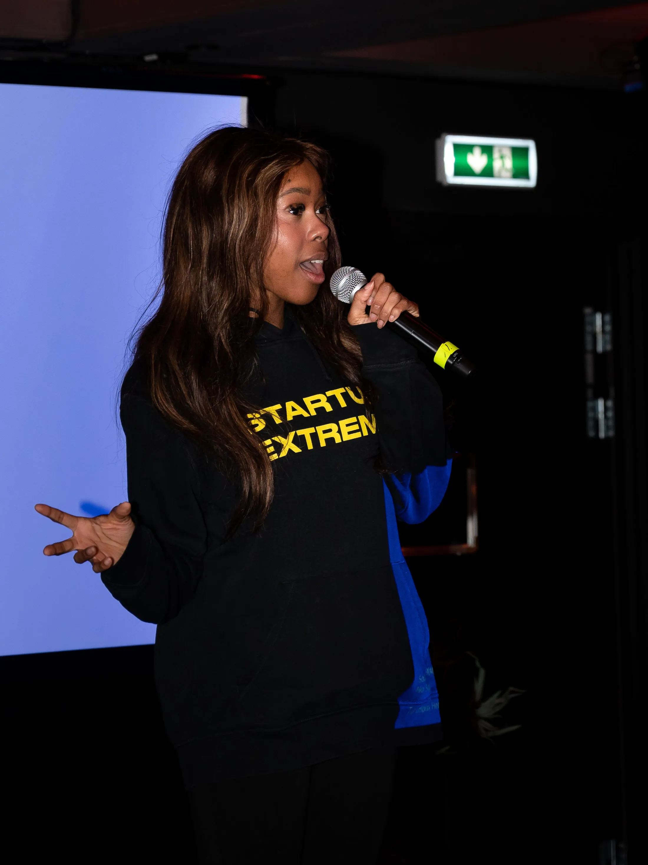 A founder in a Startup Extreme sweater pitches with a microphone on a dark stage.