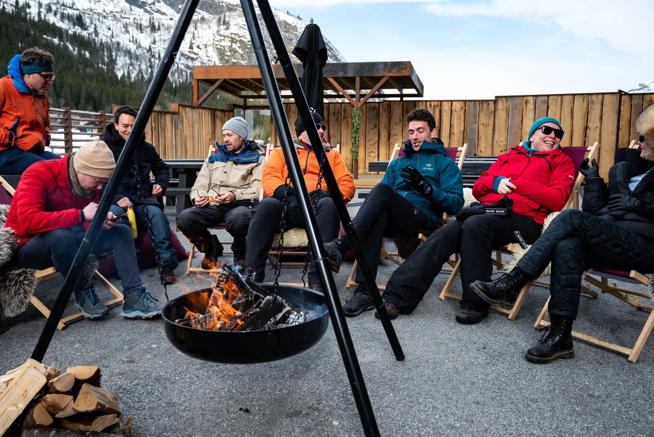 Extremers in mountain jackets laugh in deck chairs around a fire pan on a terrace, a snowy mountainside behind them.
