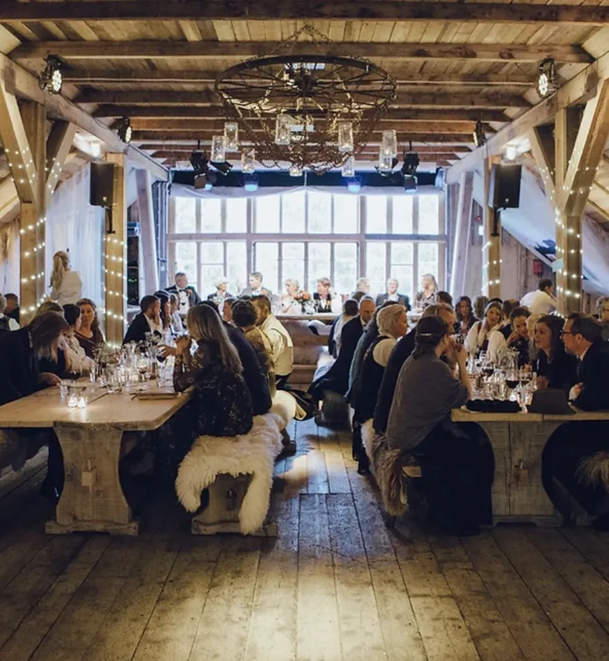 Long wooden dinner tables with sheepskins in a rustic timber hall under a chandelier.
