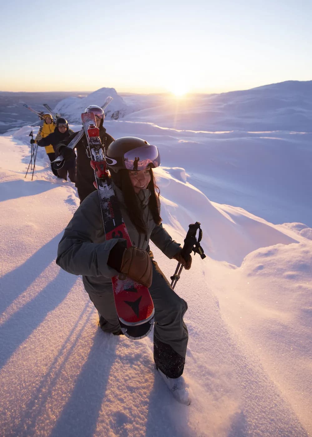 Skiers on a randonnée tour along a snowy ridge at sunset, a woman with skis on her shoulder in front.