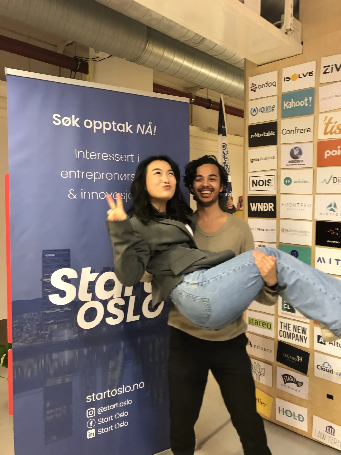 Two students laughing in front of a Start Oslo banner and a wall of startup logos, one carrying the other.